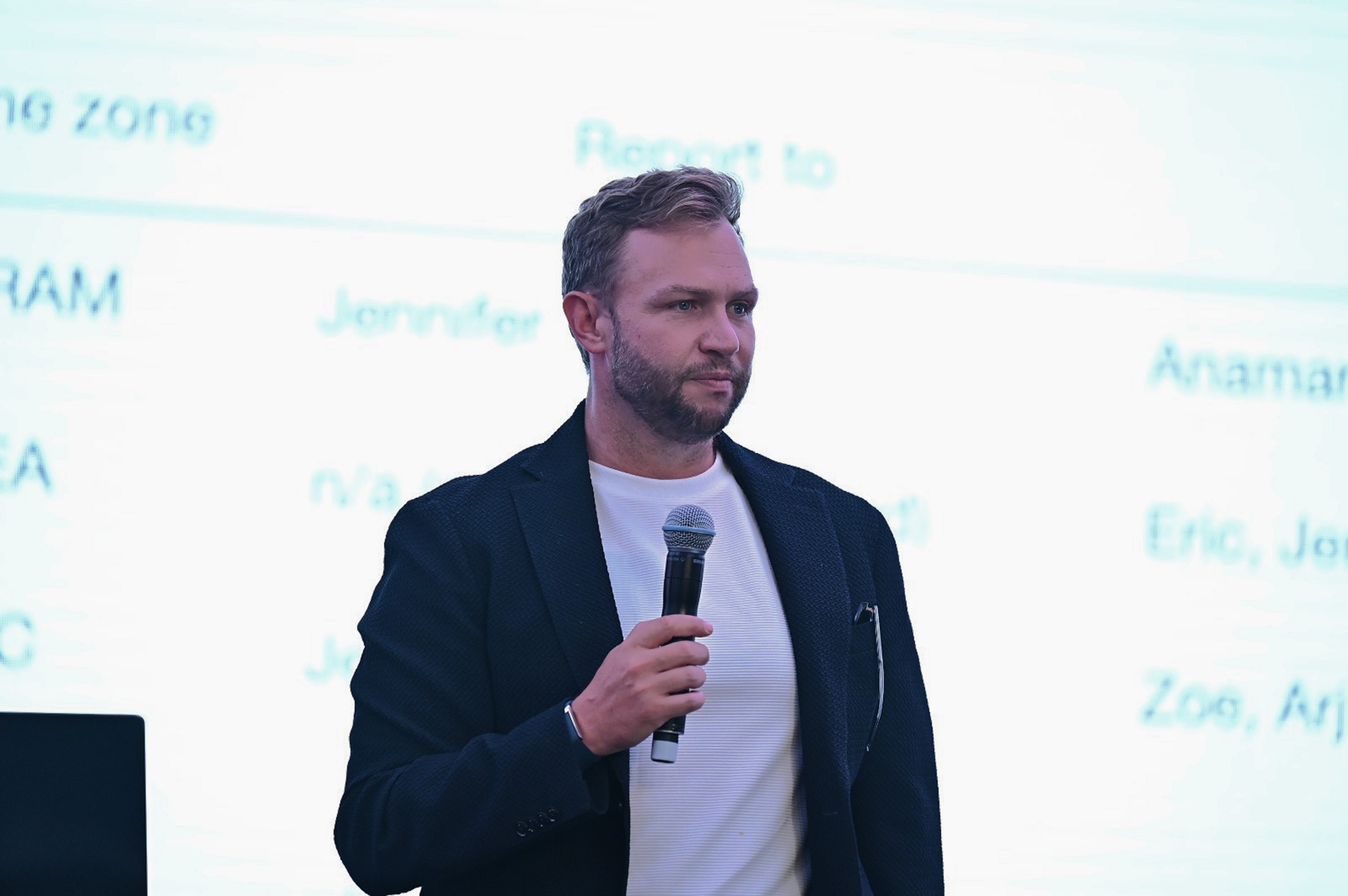 A man holding a microphone stands in front of a presentation screen during a conference.